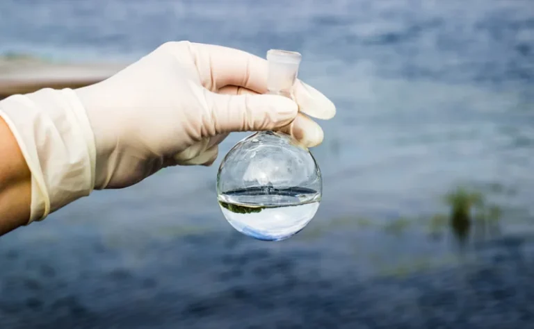 a person in white gloves holding a water sample in a beaker after water contamination occurred