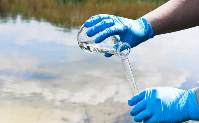 a person in blue gloves standing in front of a river pouring water from the river into a beaker for testing purposes to see if there are PFAS chemicals in the water supply